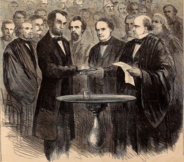 An engraving shows Abraham Lincoln standing at a small round table, placing his hand on a Bible held by another man. He is surrounded by several men in formal coats and robes, while a large crowd of observers gathers in the background to witness the ceremony.