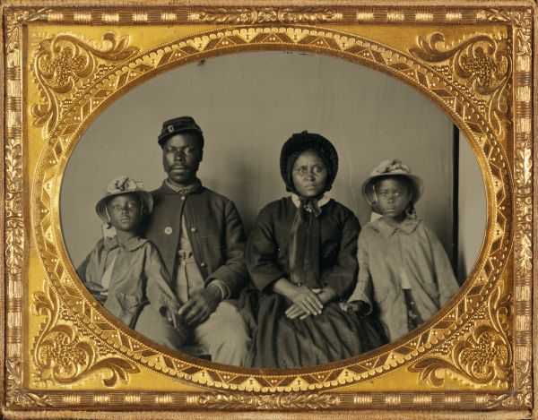 A tintype photograph shows a Black man in a Union soldier's uniform seated alongside a woman and two young children in an ornate gold frame. The family is posed formally for the portrait, with all four members looking directly toward the camera.