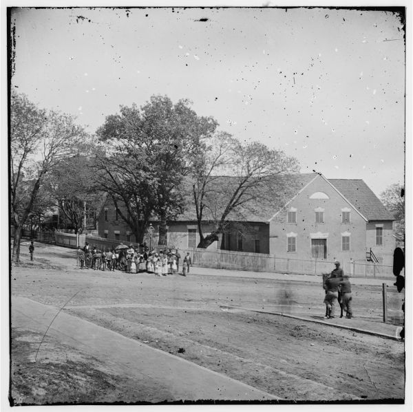 A grainy black and white photograph shows a large, two-story stone building with a gabled roof set back from a wide, dirt road. Several small figures, including a person on horseback, are scattered across the open foreground while a larger group of people stands gathered near a fence on the left side of the frame.