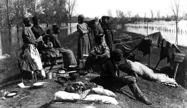A black and white photograph shows a group of Black men, women, and children displaced on a narrow strip of dry land surrounded by vast floodwaters. They are huddled together with their meager belongings, including cooking pots, bundles of bedding, and clothes draped over low wooden poles.