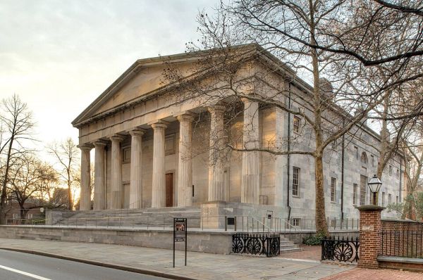 A wide-angle photograph shows the Second Bank of the United States in Philadelphia, a large Greek Revival building with a grand marble facade and a prominent row of Doric columns. The building's solid, classical architecture is captured on a late autumn or winter day, with a bare tree in the foreground and a lamppost on a brick fence.