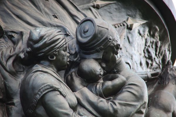 A bronze-colored relief sculpture depicts a somber close-up of a Black soldier in a military cap embracing a child. Beside them, a woman in a headwrap looks on with a mournful expression while stars and the folds of a flag are visible in the background.