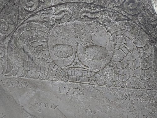 A close-up shot shows a weathered, carved gray stone with a prominent skull and crossbones motif at the top, flanked by stylized wings. Below the central design, the word 