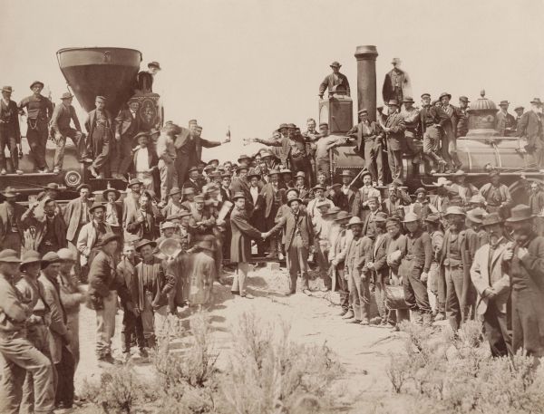 A famous photograph shows two steam locomotives facing each other as a massive crowd of workers and officials gathers to celebrate the completion of the First Transcontinental Railroad. In the center, two men lean forward to shake hands over the final golden spike driven into the tracks at Promontory Summit.