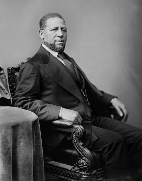 A black and white studio portrait shows Hiram Revels, a man with a beard and short hair, sitting in an ornate wooden chair and wearing a formal dark suit. He is captured from the side in a three-quarter view, looking slightly toward the camera with a neutral expression.