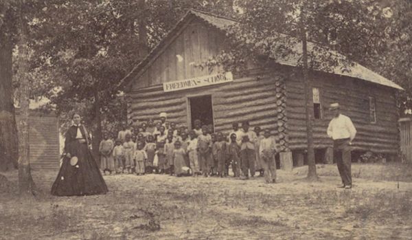 A sepia-toned photograph shows a large group of Black children standing in front of a log cabin building with a sign that reads Freedmen's School. To the left, a woman in a long dark dress stands prominently, while a man in a light-colored shirt and dark trousers stands to the right near a tree.