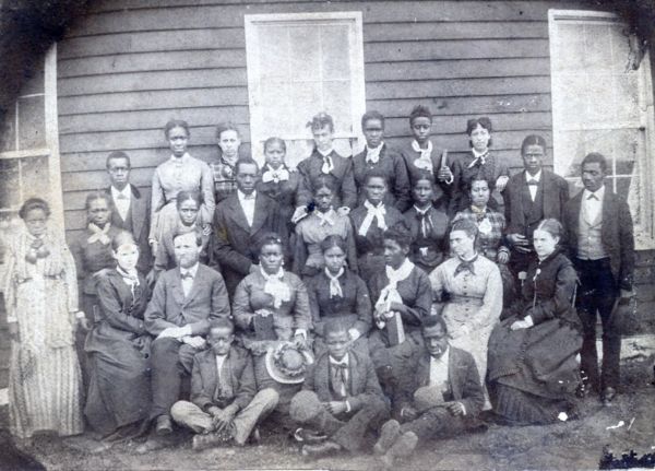 A black and white photograph shows a group of about twenty-five young men and women posed in front of a wooden building. The individuals are dressed in formal Victorian-era clothing, with many of the women wearing dark dresses with white collars and the men in dark suits and bow ties.