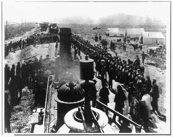 A black and white photograph taken from the top of a steam locomotive looks down upon a long line of workers standing along the railroad tracks. In the background, white tents are pitched in a flat, open field next to several horse-drawn wagons.