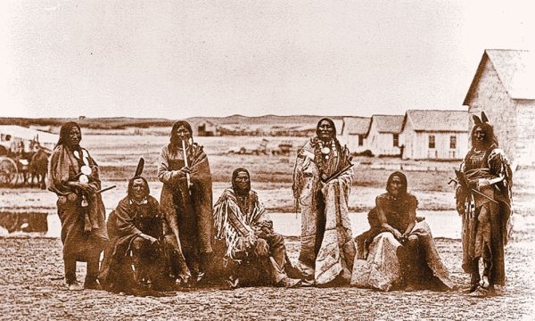 A black and white photograph shows seven Indigenous men sitting and standing in an open field in front of a row of small wooden buildings. Most of the men are wrapped in large decorative blankets, and some hold long pipes or staffs.