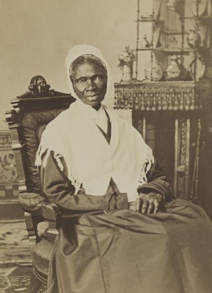 A sepia-toned photograph of Sojourner Truth, the 19th-century abolitionist and women's rights activist. She is seated in a dark, high-backed chair, wearing a dark dress, a white shawl, and a white bonnet, looking directly at the camera with a composed expression.