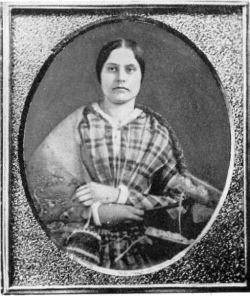 A black and white oval portrait photograph of Susan B. Anthony, the American social reformer and women's rights activist, from her younger years. She is shown seated, looking directly at the camera with a reserved expression, wearing a plaid shawl or dress and a matching shawl draped over her shoulders.