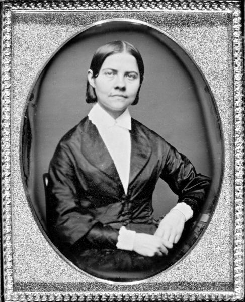 A historical black and white oval photograph of Lucy Stone, a prominent American suffragist and abolitionist. She is seated, looking directly at the camera with a reserved expression, wearing a dark fitted jacket, a white collared shirt, and a white cuff on her wrist.