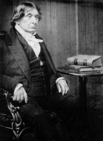 A black and white photograph of Lemuel Shaw, a prominent American jurist who served as Chief Justice of the Massachusetts Supreme Judicial Court. He is seated in a chair, wearing a dark suit and white stock, with his right hand resting on the armrest, looking off-camera with a serious expression, and two books are visible on a small table beside him.
