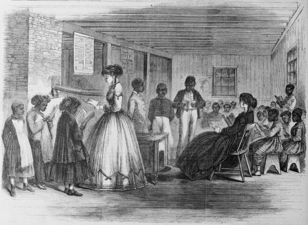 A black-and-white engraving depicts a classroom scene from the Reconstruction era where two white female teachers are instructing a group of African American children and adults. In a sparsely furnished wooden room, students stand and sit in small groups while reading intently from books and papers under the guidance of their instructors.