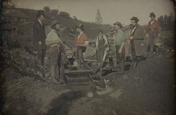A mid-19th-century daguerreotype shows a group of about eight gold miners, standing around a sluice box and other equipment at a mining site. Some of the men wear top hats and formal jackets, while others wear work clothes, suggesting a mix of laborers and proprietors at the rugged outdoor location.