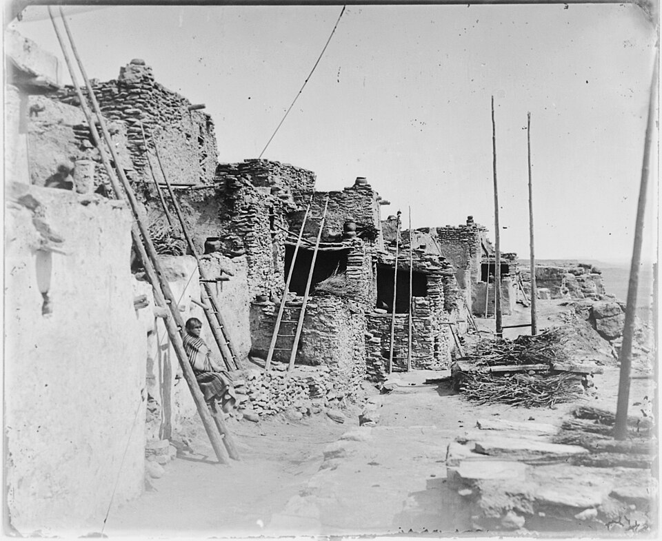 An old photo of a Hopi dwelling: a row of attached two-or-three-story apartments made of adobe brick, with a wood ladder leading from the ground to the top story. A person wearing a striped blanket around their shoulders sits on a ledge against an outer wall.