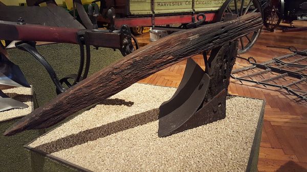 A weathered, antique plow is on display in a museum, featuring a long, thick wooden beam that serves as the handle and a heavy, curved iron blade. The plow is mounted on a pedestal covered in small, light-colored stones, with various other historical farm equipment visible in the blurry background.