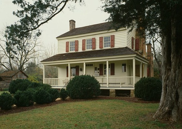 A color photograph shows a two-story house with a large front porch, a white exterior, and red shutters. The house sits on a grassy lawn with manicured bushes and is surrounded by large trees under an overcast sky.