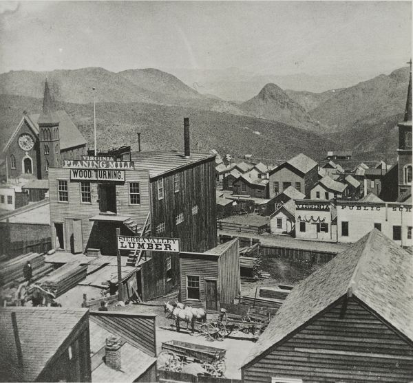 A black and white photograph shows a bustling industrial town nestled in a valley with mountains in the background. In the foreground, a large wooden building labeled Virginia Planing Mill stands next to stacks of lumber and a horse-drawn wagon.