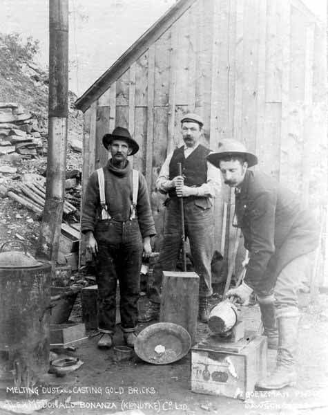 Three men stand outside a wooden shack next to a large metal furnace with a tall smokestack. One man leans over to pour molten metal from a handheld crucible into a mold, while gold-panning equipment sits on the ground nearby.