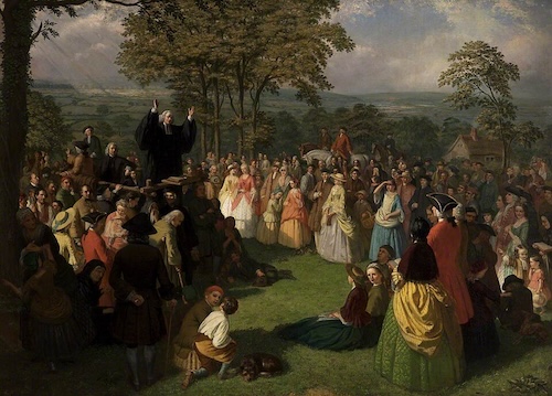 A color painting shows George Whitefield, a man in black robes, standing on a platform with his arms raised, preaching to a large crowd outdoors. The diverse crowd of men, women, and children is gathered on a grassy field surrounded by trees and rural buildings.