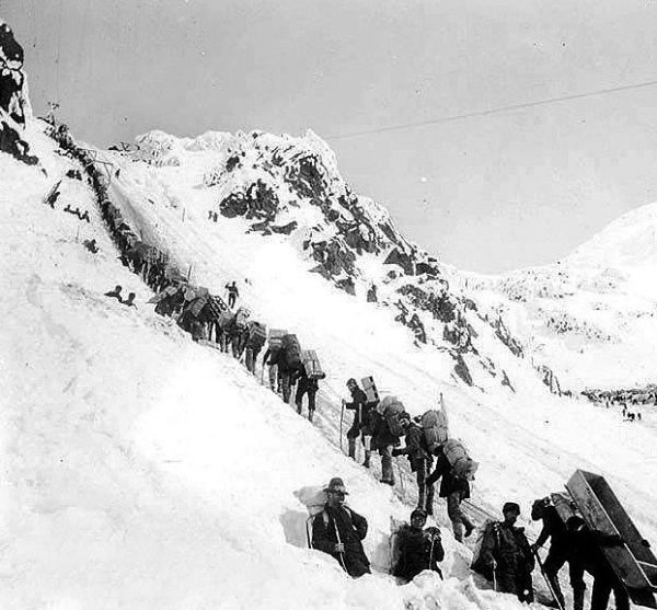 A black and white photograph shows a long line of men carrying heavy packs as they slowly climb a steep, snow-covered mountain pass. The men are dressed in thick winter clothing and are spaced out along a narrow trail carved into the deep snow.