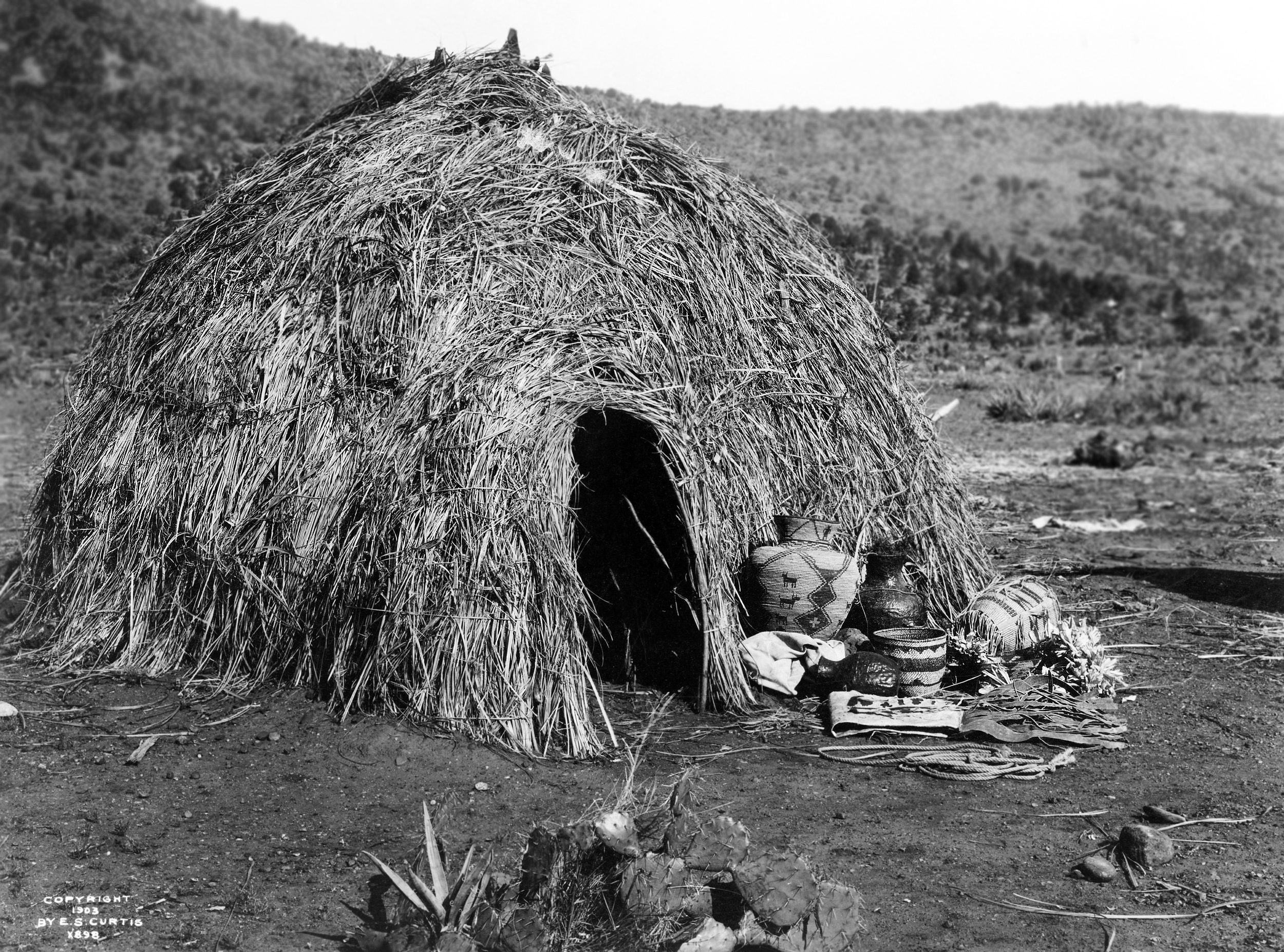 Black-and-white photograph of a dome-shaped Apache wickiup made from brush and grasses, with baskets and belongings near the entrance in a dry, open landscape.