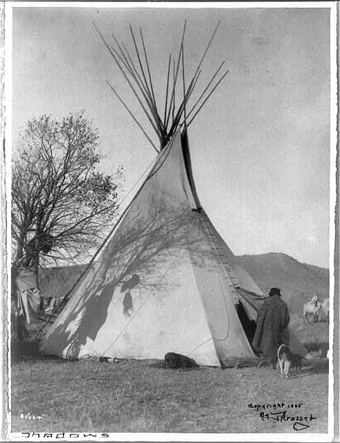 Black-and-white photograph of a tipi with tall poles extending from the top, with a person, dog, and horse nearby in an open landscape.