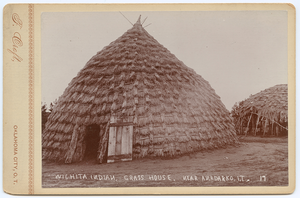 Sepia photograph of a large cone-shaped Wichita grass house covered with thatched grass, with another grass structure nearby.