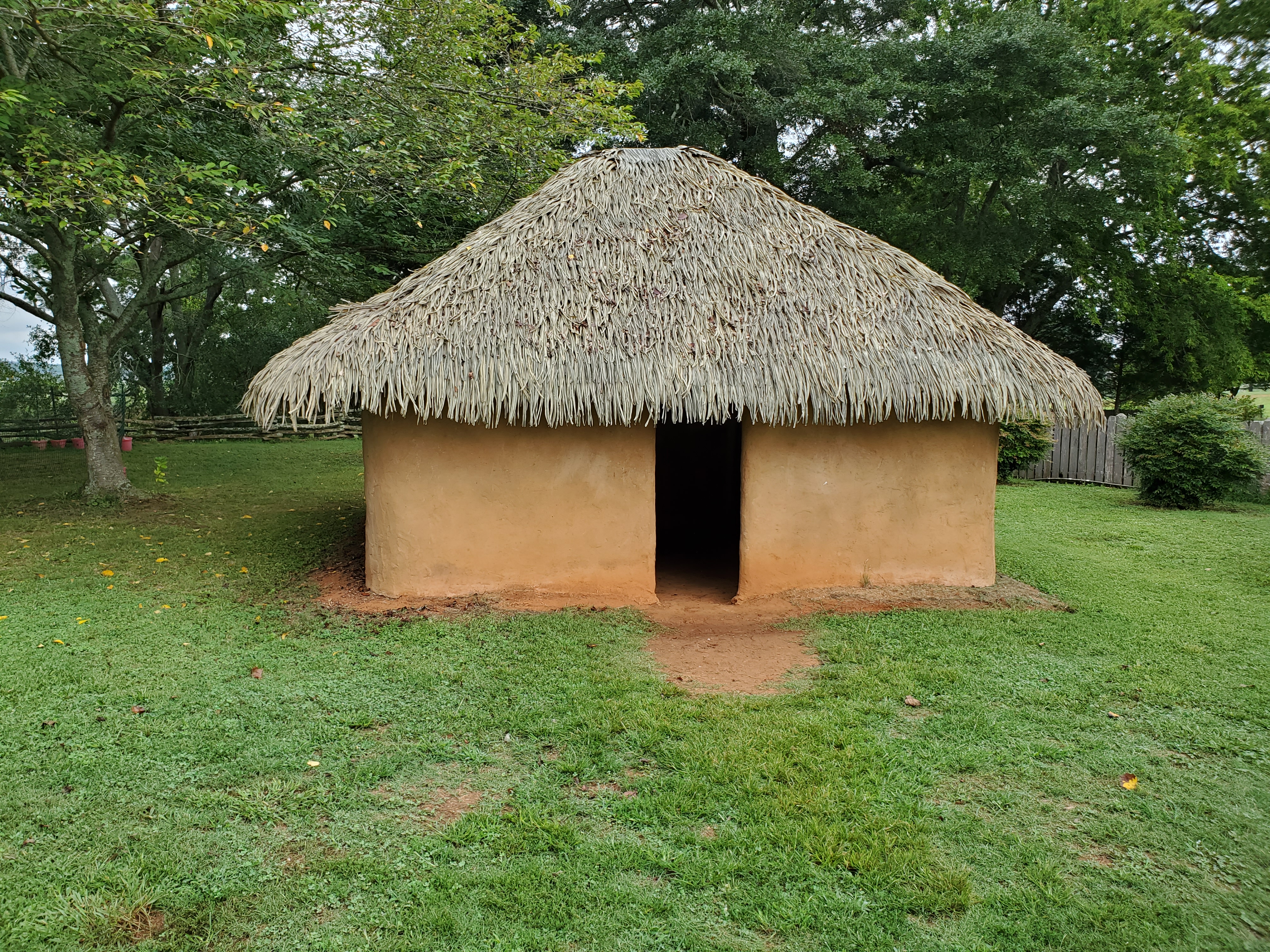 Color photograph of a reconstructed wattle and daub house with smooth clay walls, an open doorway, and a thick thatched roof, surrounded by grass and trees.