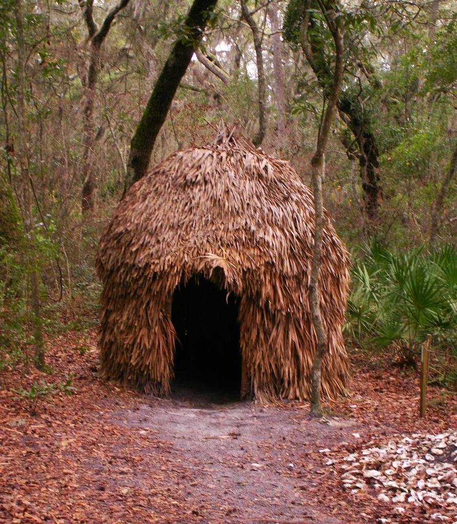 Color photograph of a reconstructed chickee-style shelter with a thatched roof and open doorway, surrounded by trees in a wooded area.