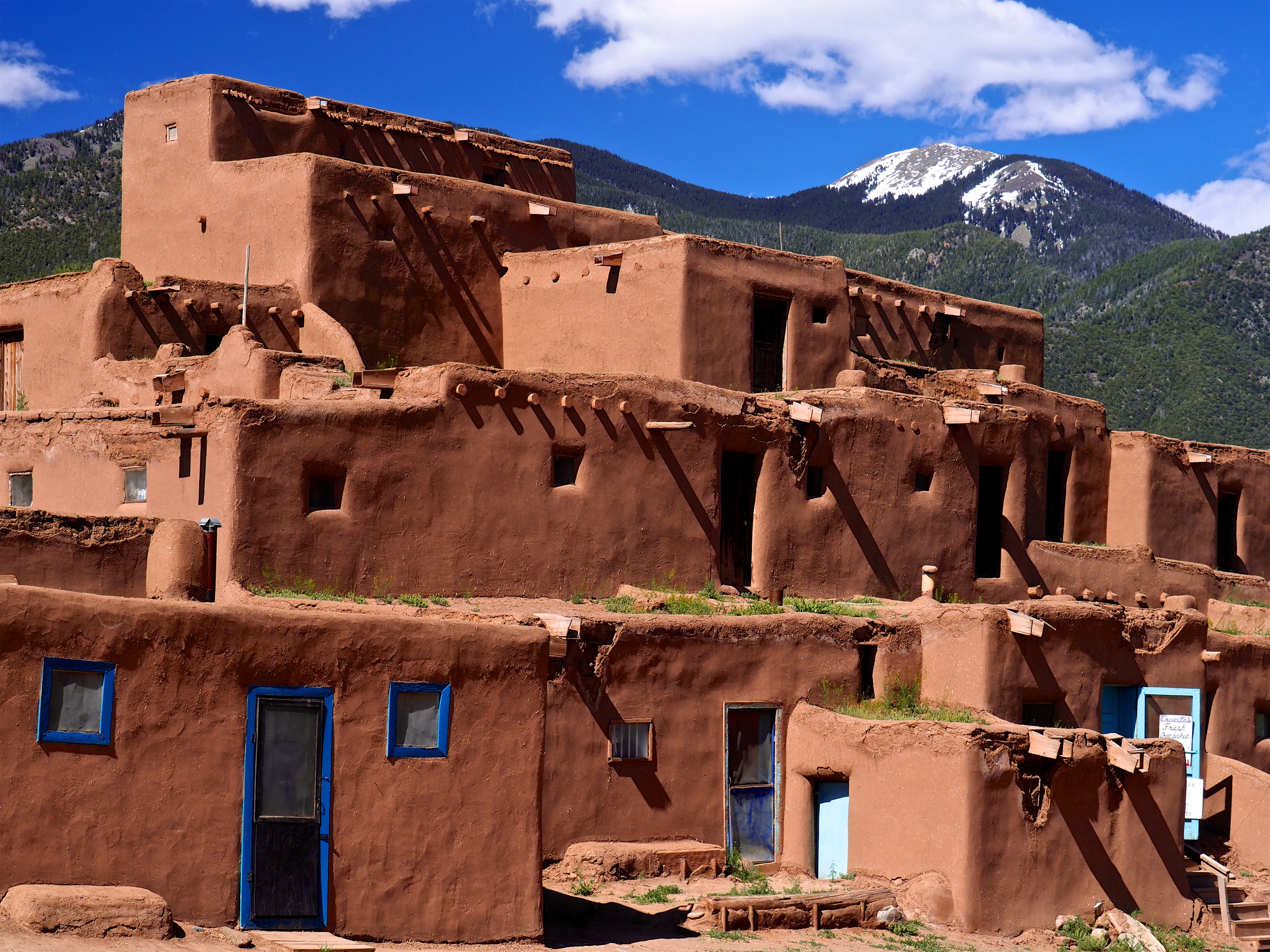 Color photograph of a multi-story adobe pueblo with earthen walls, blue doors and windows, wooden beams, and mountains in the background.