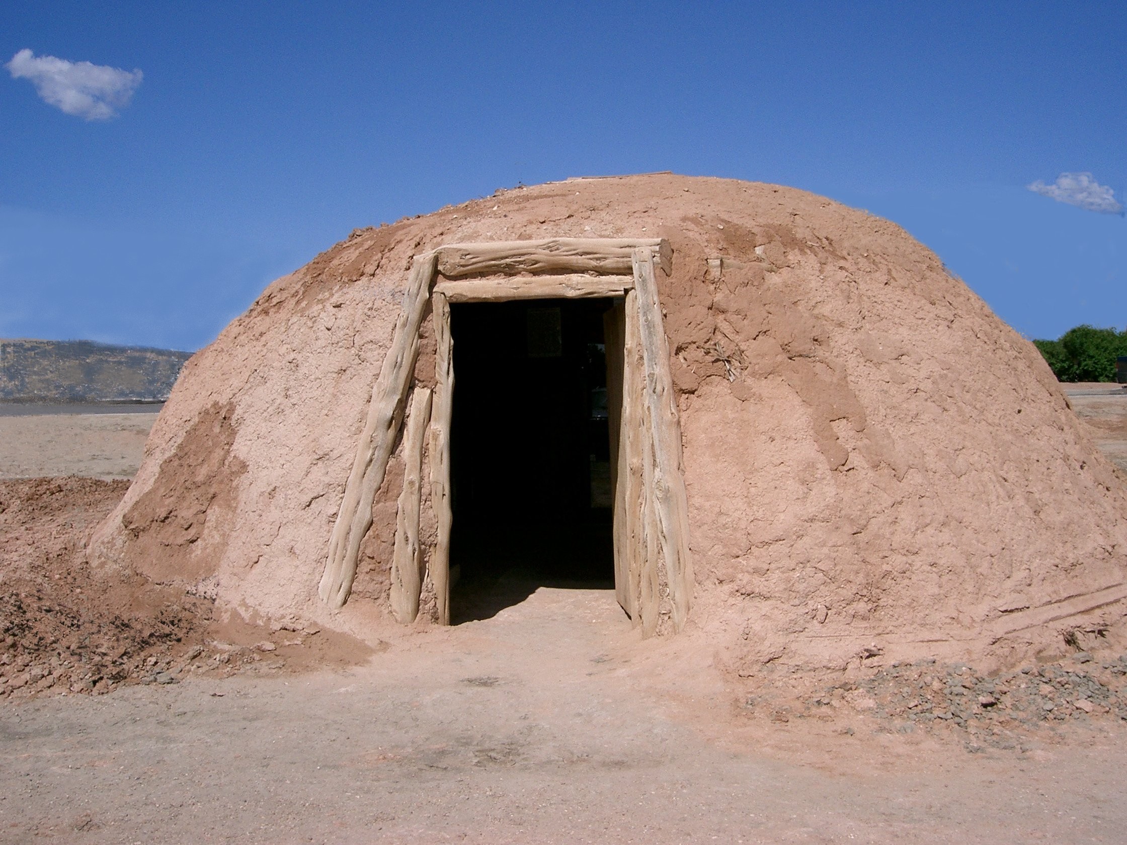 Color photograph of a reconstructed earth lodge or hogan made from packed earth, with a wooden-framed doorway and a rounded dome shape in a dry landscape