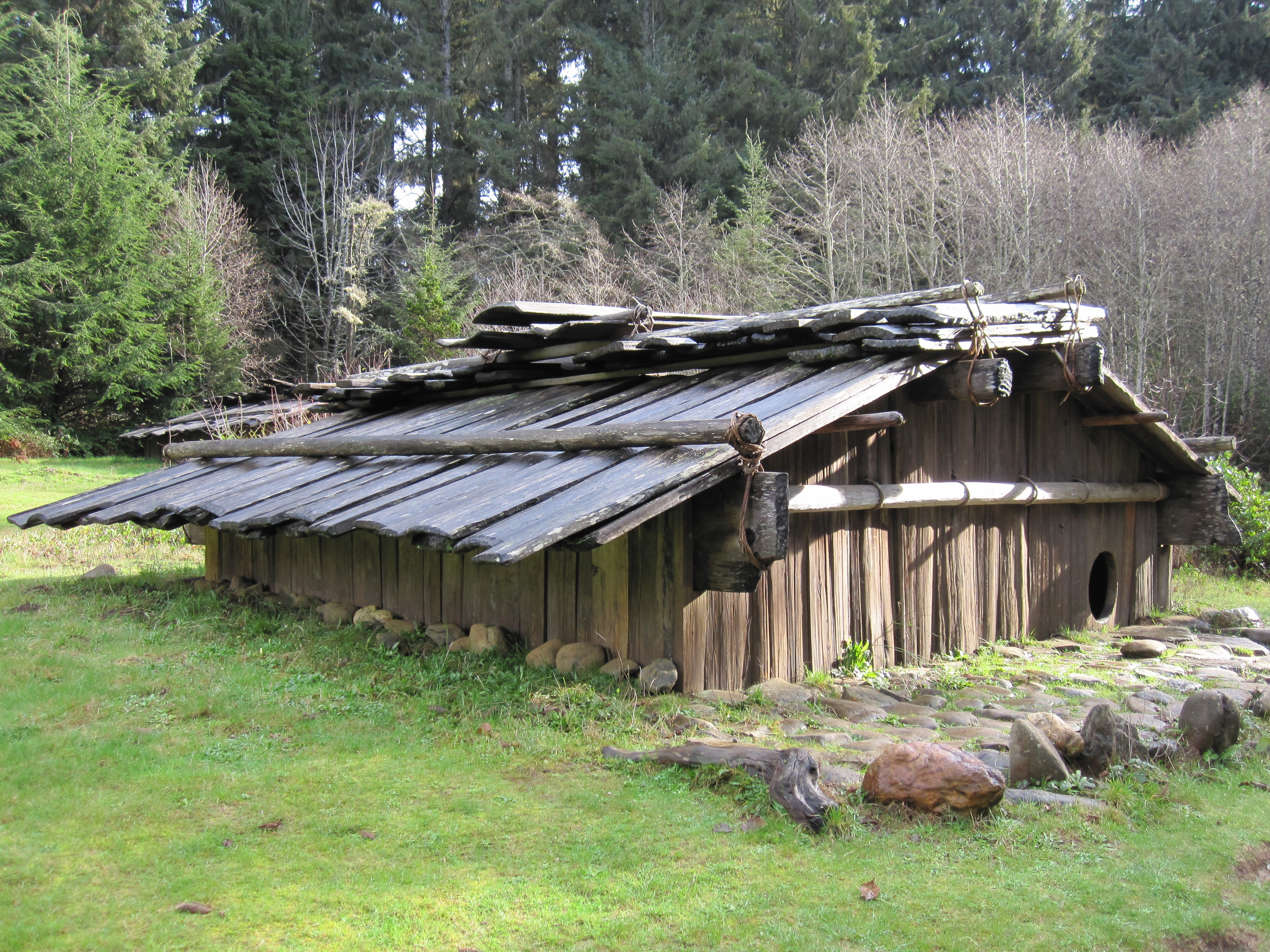 Color photograph of a reconstructed Northwest Coast plank house made from wooden boards, with a sloped plank roof, round doorway, and forest in the background.