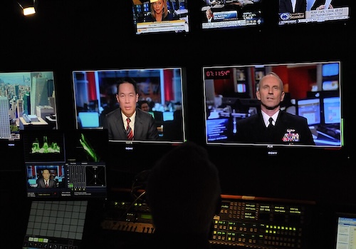 A dimly lit television control room with multiple monitors showing live news broadcasts. Two large screens display a news anchor and a uniformed military official, while smaller screens above show other news clips and financial updates. A silhouetted operator sits in front of video production equipment and control panels.