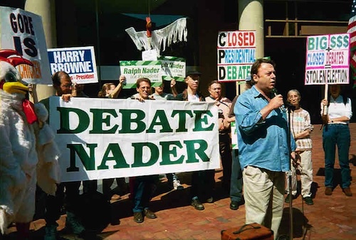 A group of protesters stands outside holding signs supporting Ralph Nader’s inclusion in a presidential debate. A large banner reads “DEBATE NADER,” while a man speaks into a microphone in front of the crowd. Other signs criticize the closed presidential debate process.