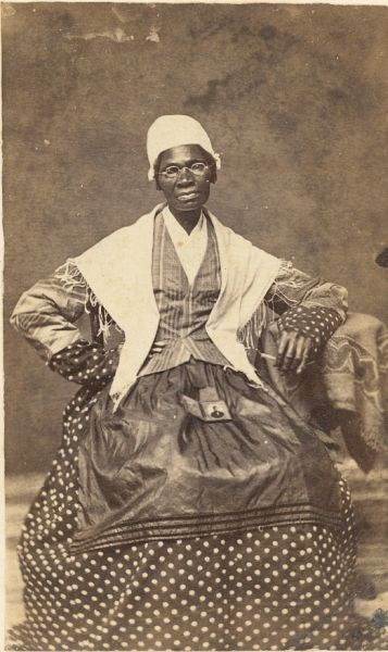 A sepia-toned carte de visite photograph of Sojourner Truth, the 19th-century abolitionist and women's rights activist, taken around 1870. She is seated, wearing a bonnet, glasses, a fringed shawl, a striped vest, and a polka-dot skirt, with a small photograph pinned to her lap.