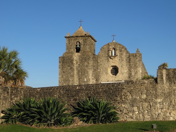 A weathered stone mission building stands tall against a clear blue sky, featuring a bell tower on the left and a domed section on the right with a statue in a niche. In the foreground, a low stone wall encloses the mission grounds, with lush green grass and agave plants visible.
