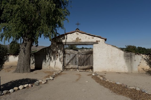 A weathered, light-colored adobe wall of a Spanish mission with a dark wooden gate in the center. Above the gate, the wall forms an archway topped with a small cross. To the left, a large, mature tree with green leaves casts a shadow. A dirt path leads up to the gate, bordered by a line of rocks on the left and wood chips on the right. The sky is clear and blue, and hills are visible in the background.