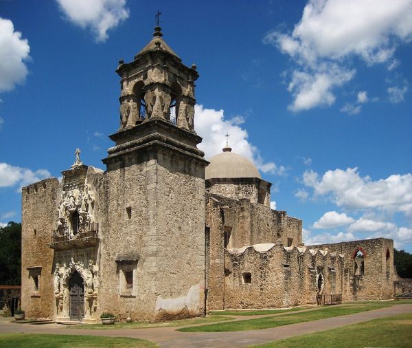 A sunlit, wide-angle photograph captures the stone structure of Mission San José in San Antonio, Texas, against a bright blue sky with white clouds. The building features a highly ornate Baroque façade, an adjoining bell tower topped with a cross, and the massive dome of the main church.