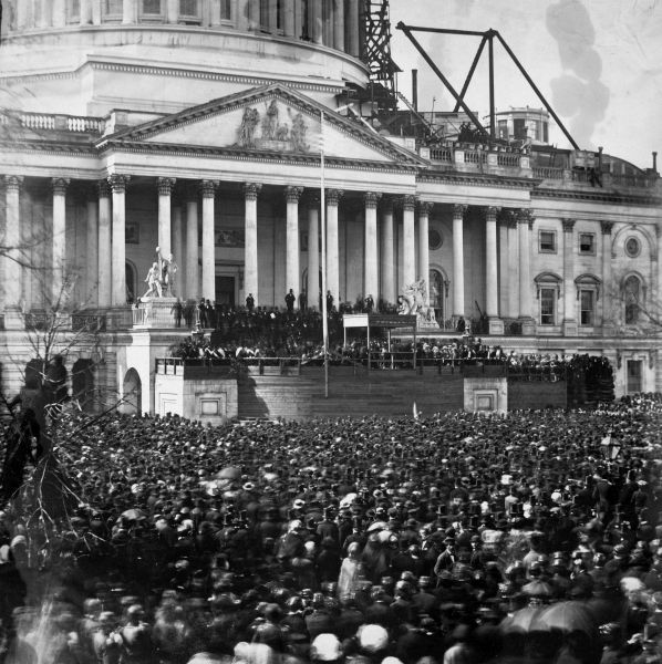A black and white photograph captures a massive crowd gathered outside the U.S. Capitol building, facing a temporary wooden platform built over the steps. People stand on the platform, likely dignitaries or officials, with a flagpole centered in front of the Capitol's columned portico, suggesting a major public event such as an inauguration.