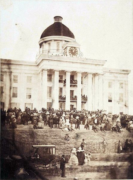 A sepia-toned photograph shows a large crowd gathered outside a grand, white Capitol building with a central dome and clock. The building has a tall portico supported by white columns, and people are visible on the balconies and steps, with the crowd extending into the foreground where a carriage is parked.