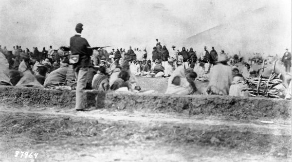 A black and white photograph shows a large crowd of Indigenous people sitting on the ground in a wide, open field. In the foreground, a U.S. soldier stands with his back to the camera, holding a rifle and watching over the group.