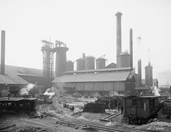 A black and white photograph shows a massive industrial iron works complex with several tall smokestacks and large cylindrical furnaces. In the foreground, steam rises from the ground near railroad tracks where a small train car and a few workers are visible among piles of industrial materials.
