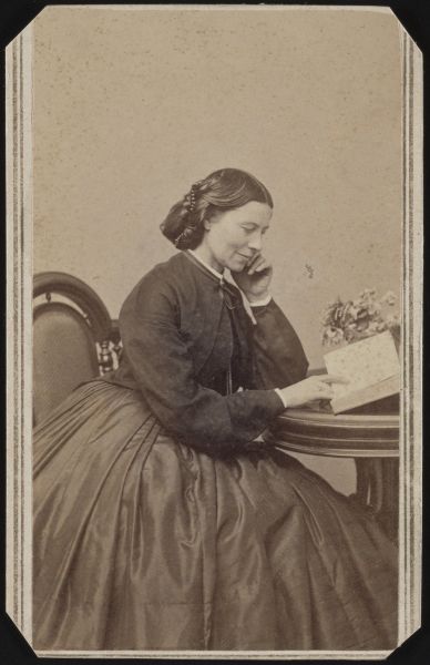  In this sepia-toned portrait, Clara Barton is shown from the side sitting at a wooden table and looking down at an open book. She wears a dark, long-sleeved dress with a full skirt and has her dark hair pulled back into an elegant style with braided details.