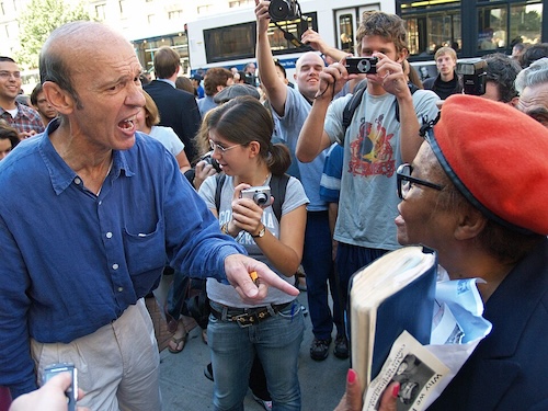 A man points while speaking during a public disagreement as people nearby watch and record with cameras.