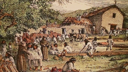 A colorized historical illustration depicts a busy Spanish mission construction site. Indigenous people, dressed in simple tunics, are engaged in various tasks: some are working with wood, others are carrying materials, and a group on the left appears to be interacting with Franciscan friars in brown robes. In the background, stone and adobe buildings are under construction or being repaired, with workers on rooftops and scaffolding. Livestock, possibly oxen, are also visible. The scene is set outdoors with trees on the left and hills in the distance.