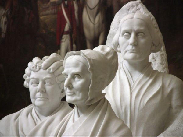 A white marble monument features the detailed portrait busts of three prominent women from the American suffrage movement: Elizabeth Cady Stanton, Lucretia Mott, and Susan B. Anthony. The sculptures are set against a dark, blurred background that highlights their stoic expressions and period-accurate clothing.