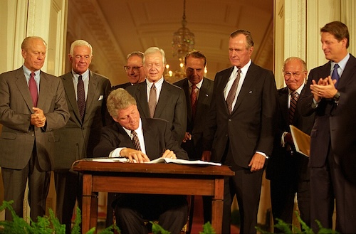 President Bill Clinton signs a document at a formal ceremony while former presidents and officials stand behind him.